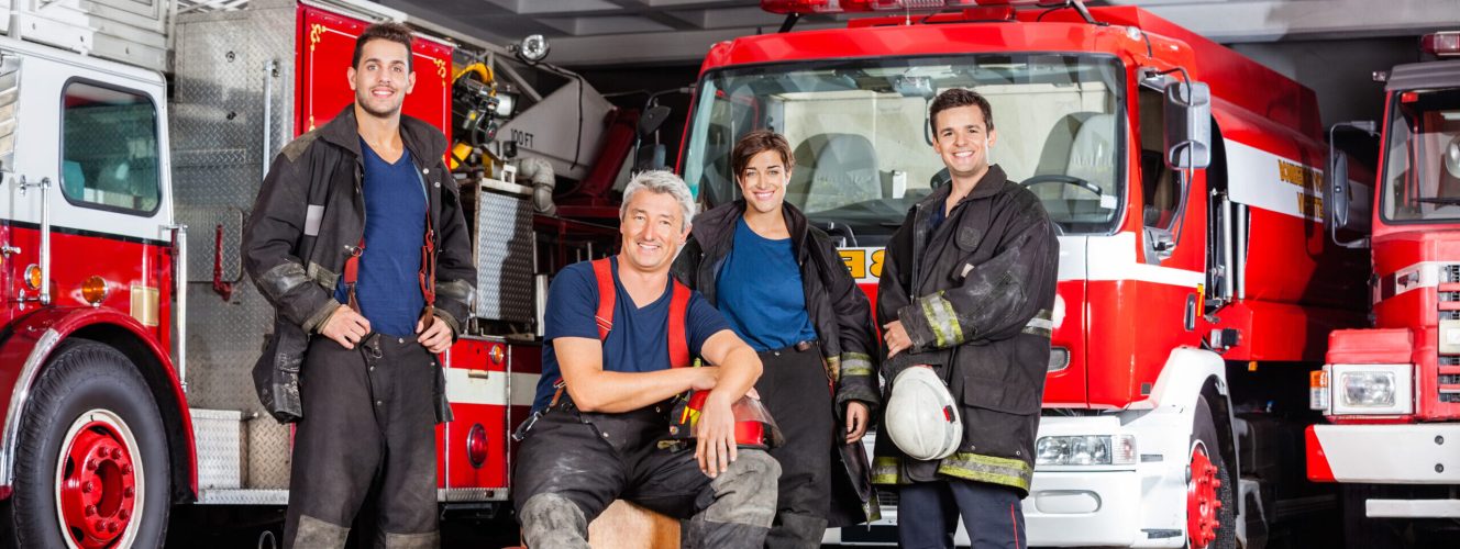 A group of safe and strong firefighters in the station with firetrucks behind them